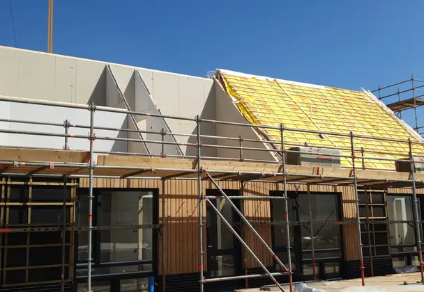 Gebouw in aanbouw met steigerwerk, houten gevel en montage van een dak met gele isolatie.