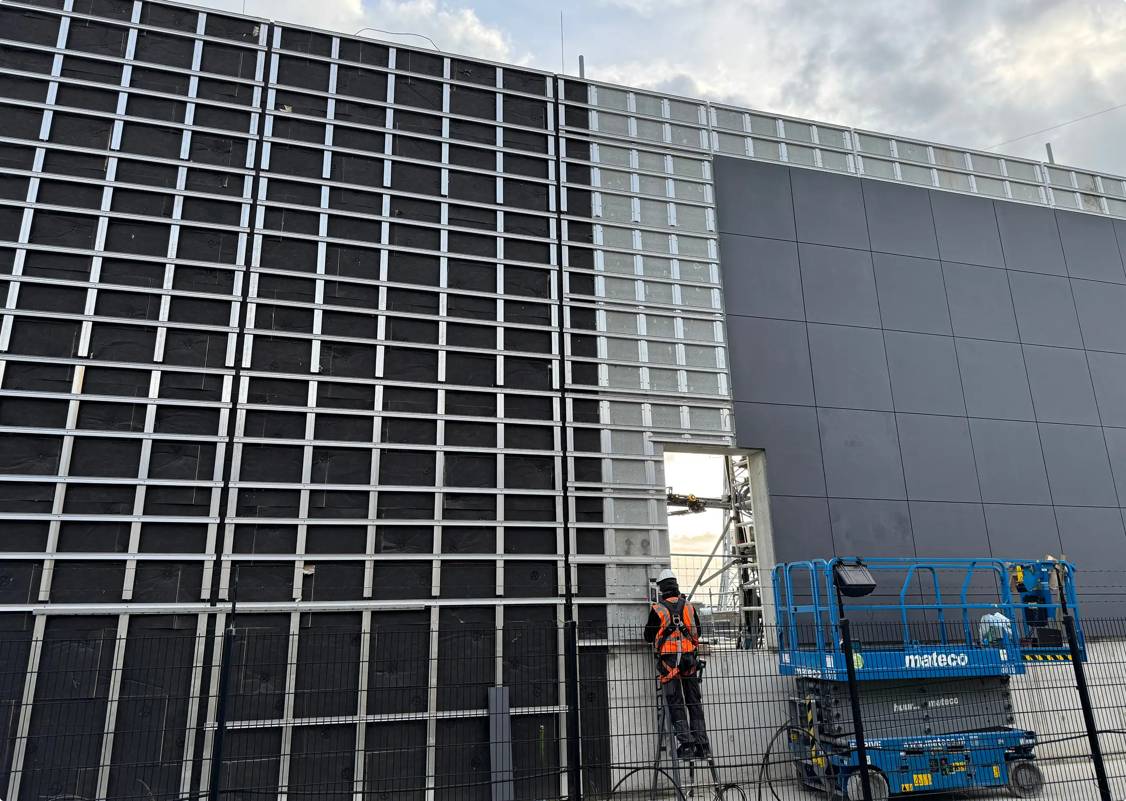 Montage van een geventileerde gevel; een werknemer op een hoogwerker bevestigt gevelpanelen.