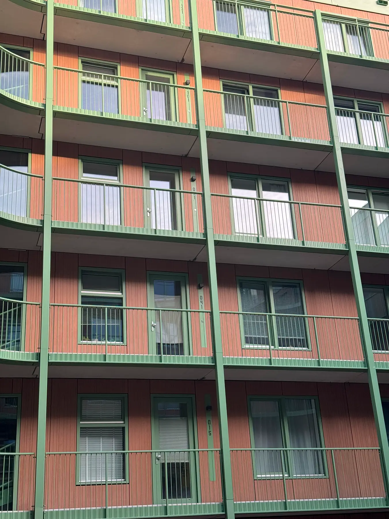 Gevel van een modern woongebouw met oranje gevelbekleding en groene balkons, raam- en deurkozijnen.