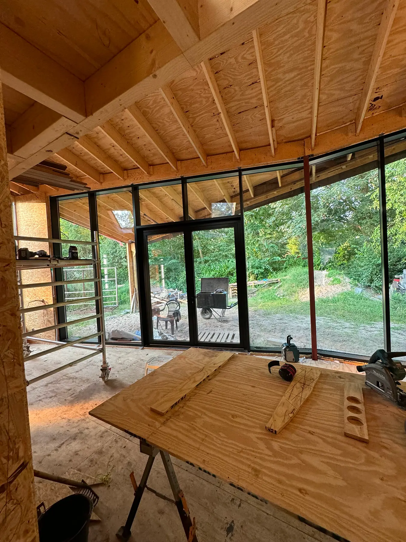 Huis in aanbouw met houten plafond, werkbank en grote glazen schuifdeuren met uitzicht op de tuin.